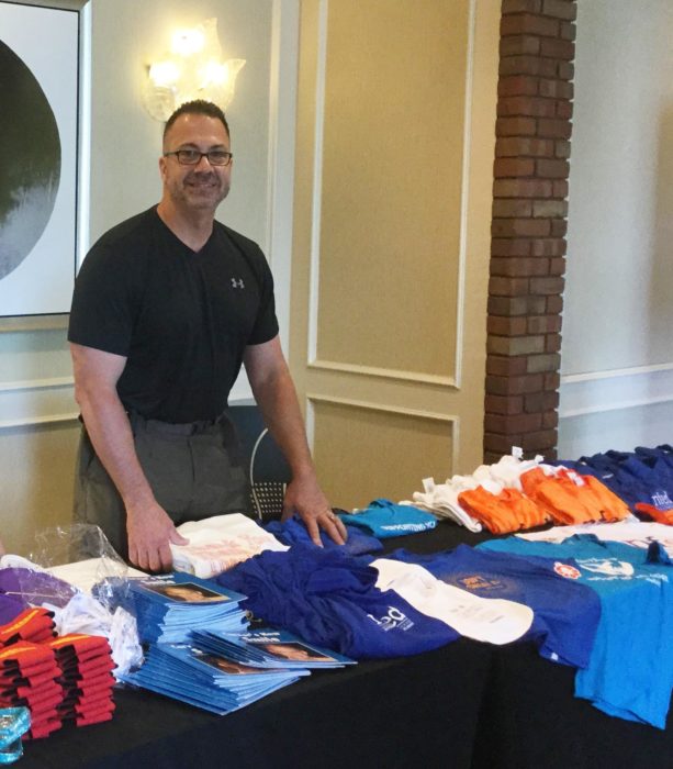 Paul volunteers and stands behind a table filled with NFED t-shirts, books, and other items for sale at the Family Conference.