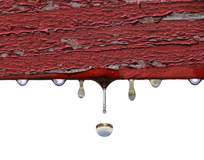 A picture of a gray piece of wood with red paint peeling off and several rain drops falling off the bottom. In the reflection of the rain drops you can see trees and sky.