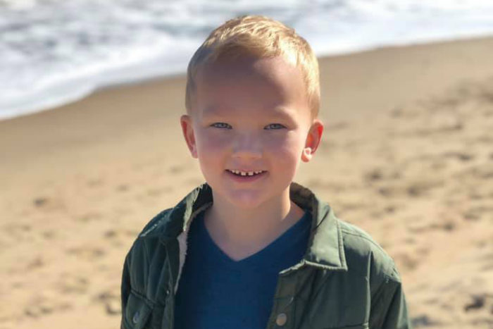 Boy standing on the beach with the ocean behind him. He's wearing a jacket and smiling.