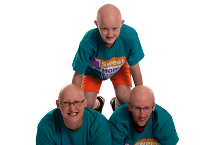 A tween boy with AEC syndrome kneels on the backs of an adult woman and adult man also with AEC syndrome.  They all have matching teal shirts and are smiling. 
