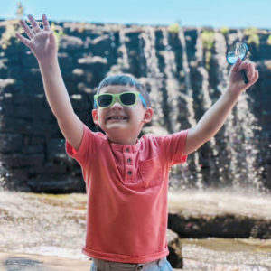 Keller is wearing a read shirt and standing in front of a waterfall raising his arms into the air. He has blue hair and sunglasses on, smiling.