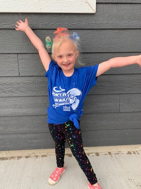 Ave is wearing her Don't Sweat It Walk shirt with her hands held above her head. Her hair is in three tiny ponytails which are painted green, red and blue.
