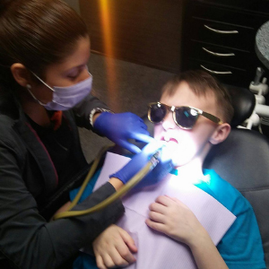 A dentist works on the mouth of a boy who's in a dental chair. The boy is wearing sunglasses and a blue shirt.