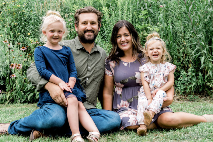Tyler and Erin Nawrocki sit in the grass with a garden behind them. Their daughters, Ava and Aliana, sit on their laps.