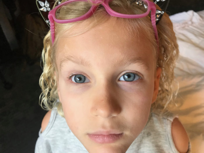 Lily is looking into the camera. She's wearing a gray shirt and has blonde curls with a pink glasses on her head. 
