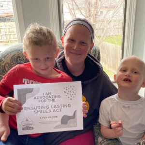 A mom has a daughter on her lap and her son at her side. The daughter is holding a sign that says, "I advocated for the Ensuring Lasting Smiles Act."
