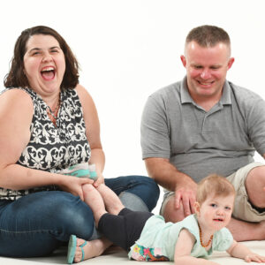 Mom and dad sitting on the floor laughing and smiling. Little girl is laying down in front of them smiling.
