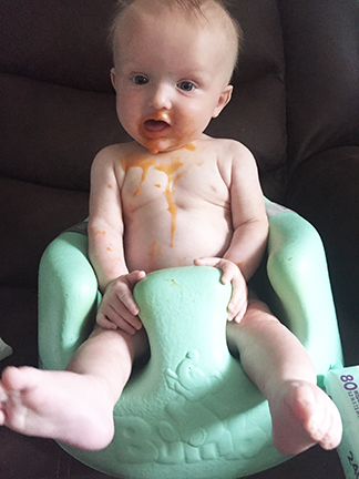 A baby is sitting in a high chair without a shirt on. He has baby food on his face and dribbling down his chest.