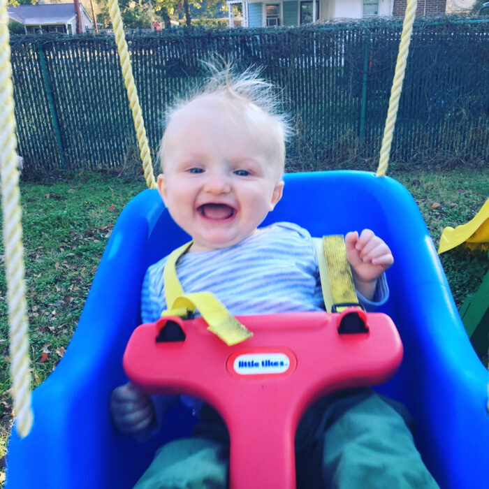A baby is smiling and in a swing that is red and blue. The baby has sparse, blonde hair that is sticking up.