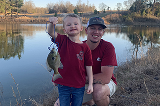 A boy affected by hypohidrotic ectodermal dysplasia holds up a string with a fish on it. His dad squats down behind him. They are in front of a lake.