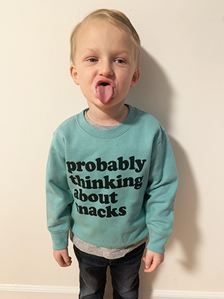 A toddler boy with blonde hair is sticking out his tongue. He's wearing a teal shirt that says, "probably thinking about snacks."