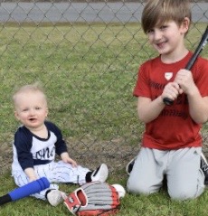 Beau and his brother are wearing baseball uniforms and sitting on the ground at a baseball field. One boy is holding a baseball bat. There's a bat and glove on the ground.