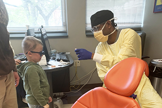 A dentist in scrubs and a mask holds a pair of dentures which he's showing a four-year-old boy.