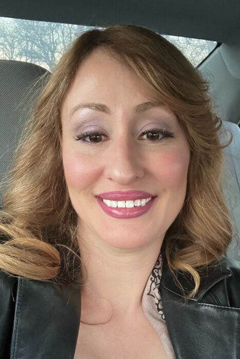 A woman with medium length medium brown hair smiles. She's wearing a black jacket.