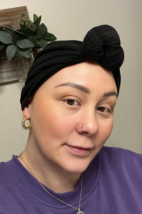 This is a headshot of a woman who is wearing a purple t-shirt and a black scarf around her head. 