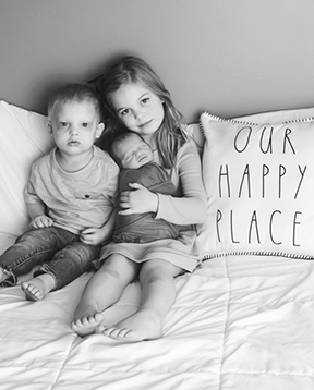 Three children sit on a bed with a white bedspread. A pillow says, Our Happy Place." A young girl holds a small baby wrapped in a blanket. A toddler boy is next to them.