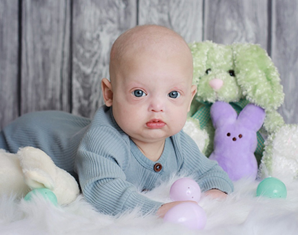 A baby lies on his belly on a white rug surrounded by Easter eggs and stuffed rabbits. He's wearing a blue sleeper.