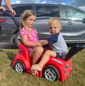 A young girl and her toddler brother sit on red plastic car while someone pushes the handle.