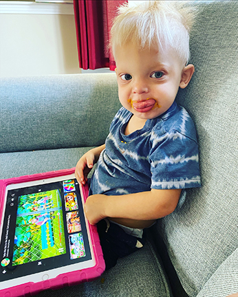 A toddler with sparse white hair sits on a couch playing game on his tablet. He has a blue shirt with white stripes. He has orange baby food on his chin.