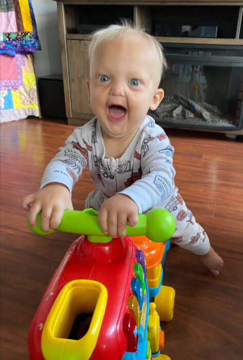 A toddler boy with sparse blonde hair and big blue eyes is riding a toddler train. He's wearing pajamas and has an excited look on his face.