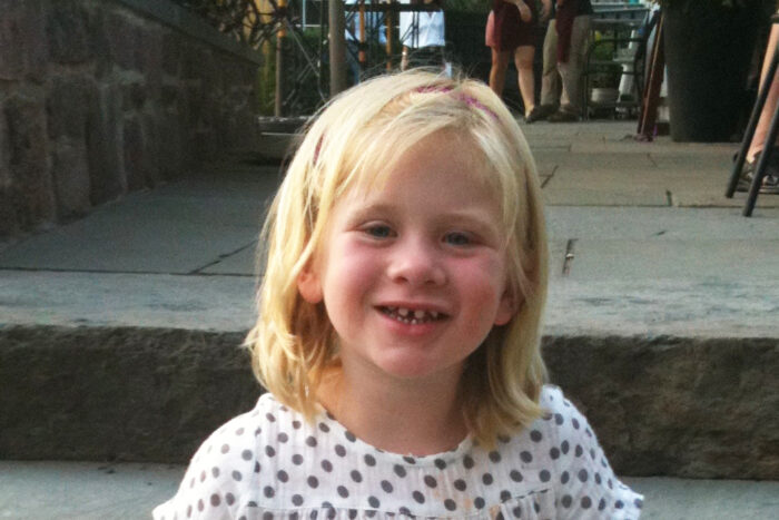 A girl is sitting on a step in an urban setting. She's wearing a white shirt with gray polka dots. She has blonde hair and is smiling. She is missing several teeth on top due to ectodermal dysplasia.