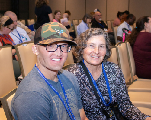 A man with a camo hat and black glasses is seated next to his mother in a conference room.