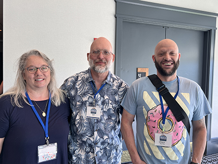 This is a picture of a woman and two men standing side by side who are family. The woman has long gray hair, glasses and is wearing a conference lanyard. The man in the middle is bald, wearing glasses and has a gray beard. He's wearing a blue shirt with flowers. The second man is also bald and has a dark brown beard. He's wearing a blue t-shirt a pink donut on it. 