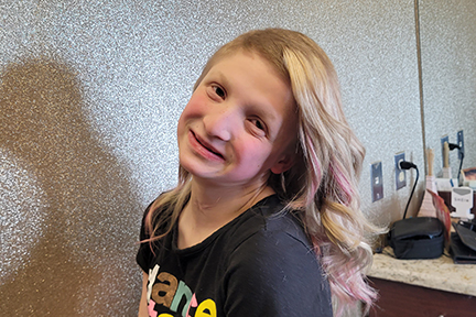 A 12-year-old girl with a p63 variation genetic disorder is wearing a black t-shirt. She has long blonde hair with pink streaks.