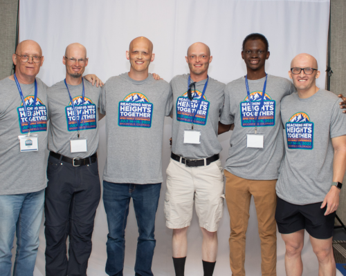 Six men of varying ages stand shoulder to shoulder, all wearing the same gray NFED Family Conference t-shirt. 