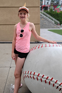 A 12-year-old girl is standing next to giant baseball. She's wearing a pink tank top, black shorts and has a unicorn baseball hat on.
