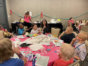 Dr. Marik holds her index finger to her lips as a group of children sitting at a round table do the same.