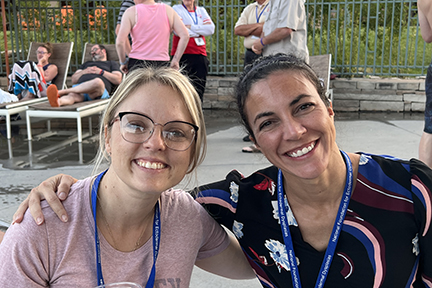 Two women are sitting on a patio with their arms around each other, smiling. One woman is blonde and wearing glasses. The other woman has dark hair pulled back.