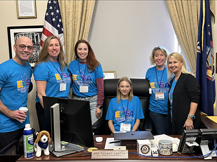 Nicole is sitting in a legislator's chair with five adult advocates all wearing the same shirt standing around the desk. A United States flag is hanging in the background.