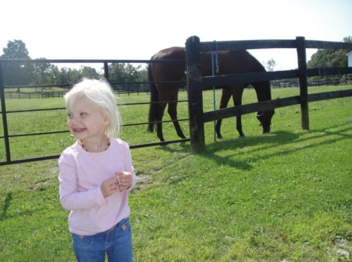 Nicole, a toddler girl with white blonde hair is smiling. She's missing several teeth. She's standing in a pasture with a fence and horse in the background.