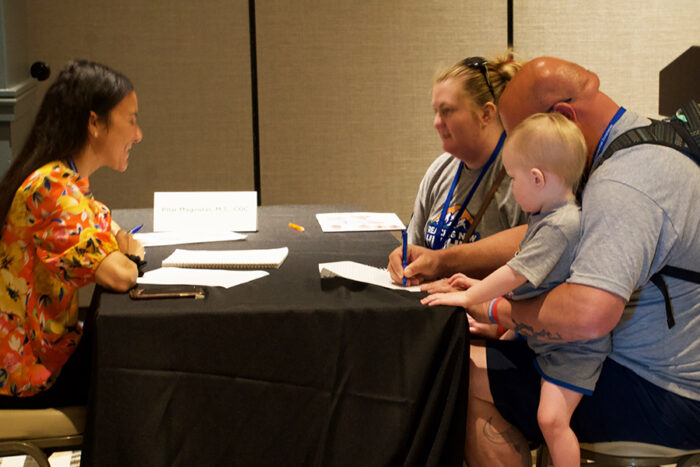 Genetic counselor, Pilar Magoulas, sits at a conference table with a black tablecloth across from a mom, dad, and their toddler son. The dad is filling out paperwork.