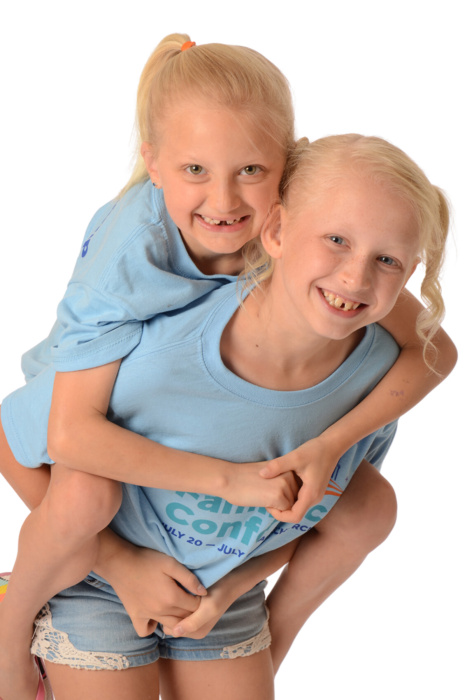A young girl with a blonde ponytail and missing teeth is on the back of another young girl who has two ponytails. They are wearing matching shirts.
