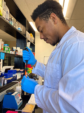 A student in a white lab coat with dark hair and facial hair injects a liquid into a tube. in a research lab.