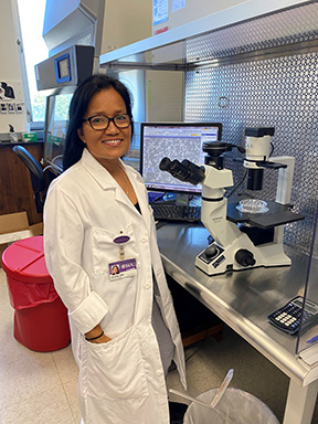 A woman with long dark hair and glasses in a white lab coat stands next to a microscope in a research laboratory.